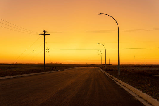 Orange sunset haze over a street with power and light poles leading to a grassy field, capturing the warm glow of the evening light and the tranquil transition from urban to nature.